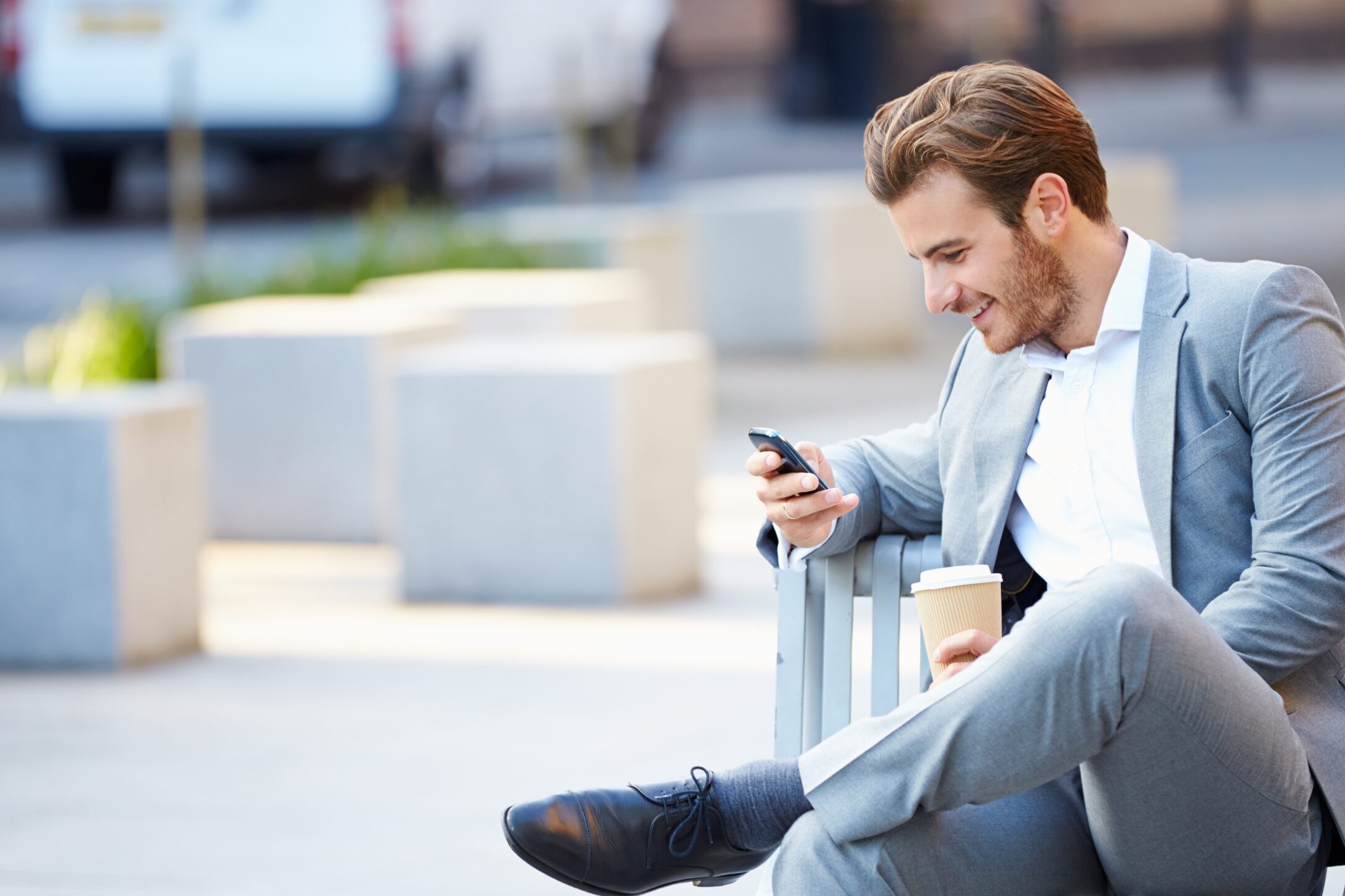 Young businessman sitting on park bench looking at phone and smiling.