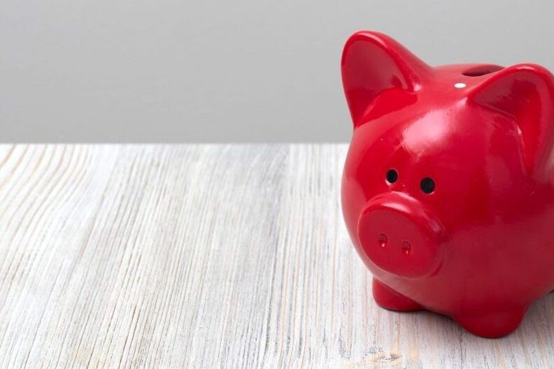 Close up of small red piggy bank sitting on wood table in front of blank grey wall.